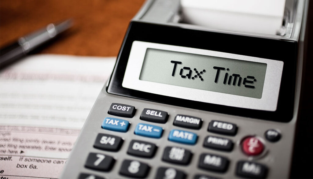 Image of a desk surface scattered with tax forms. A large calculator sits atop the papers with the words Tax Time on the display.