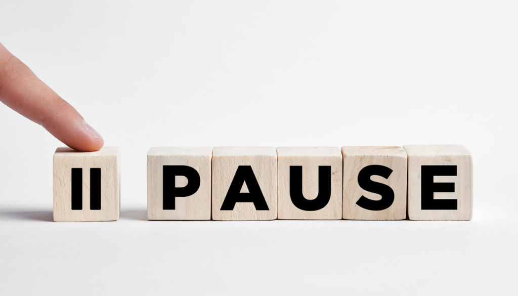 Six wooden blocks are lined up in a row. A finger from out of frame presses down on top of one that has a pause button symbol on the front face. The other blocks each have a single letter on the front face spelling out Pause.