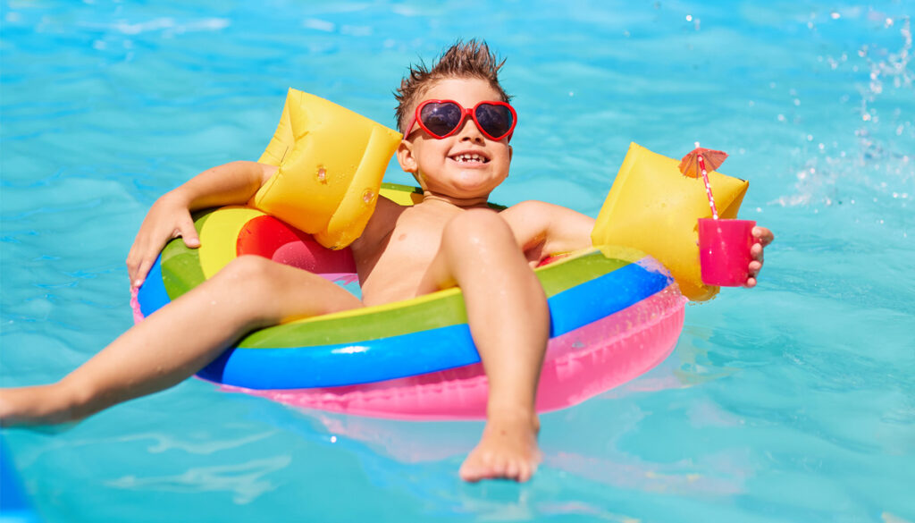 A young boy wearing red heart shaped sunglasses floats in a pool on an inner tube while wearing floaties.